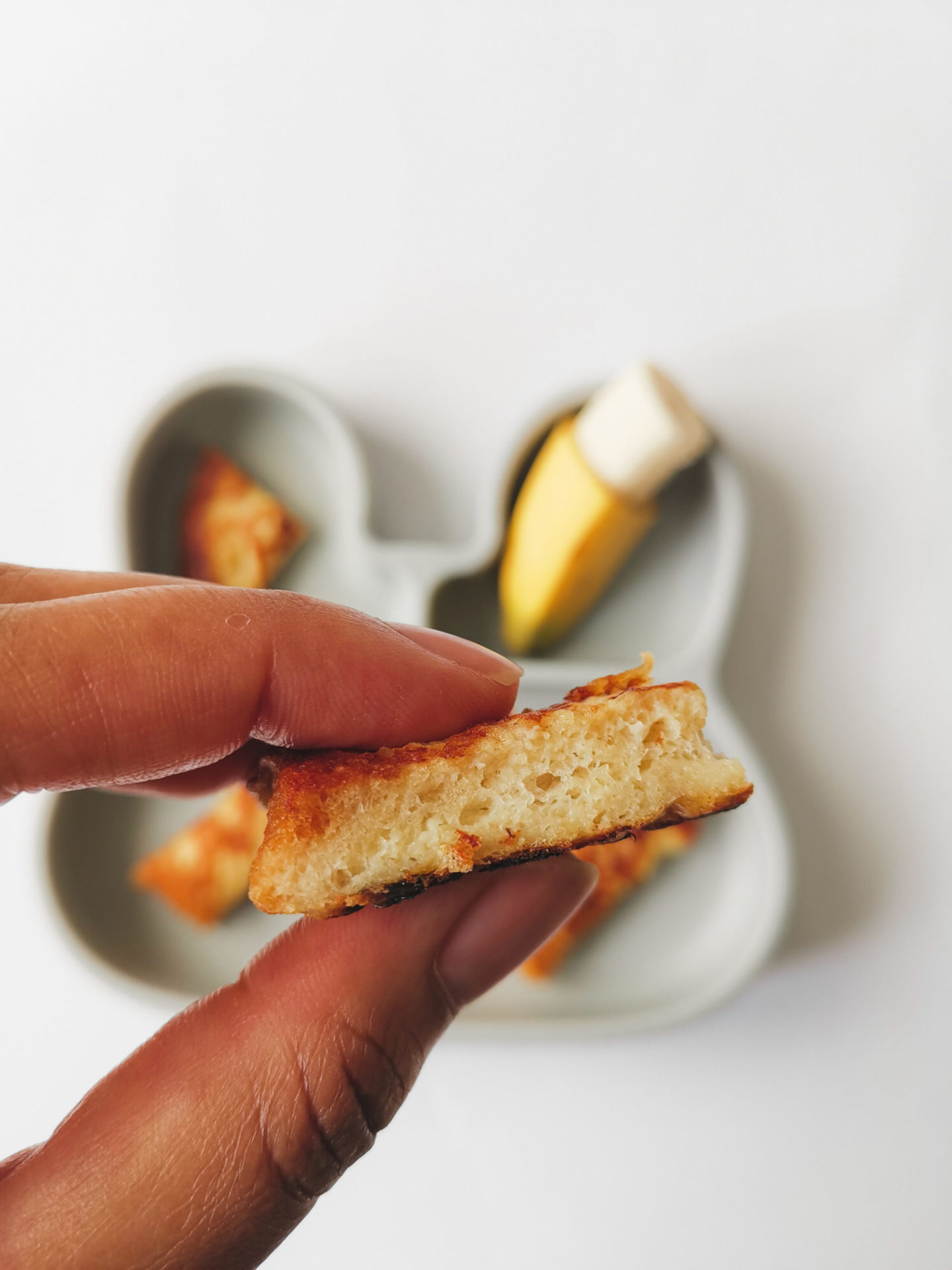 Baby French Toast: Hand hält Stück French Toast, Teller im Hintergrund